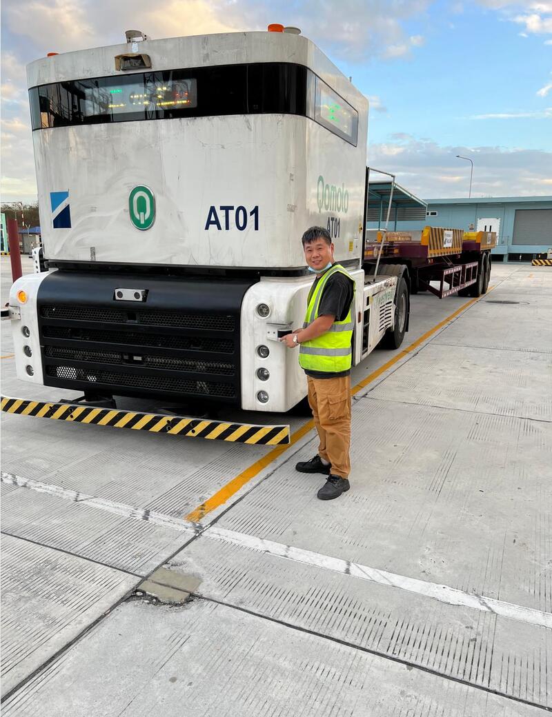 A technician stands beside an autonomous electric truck during outdoor testing at an industrial logistics facility.