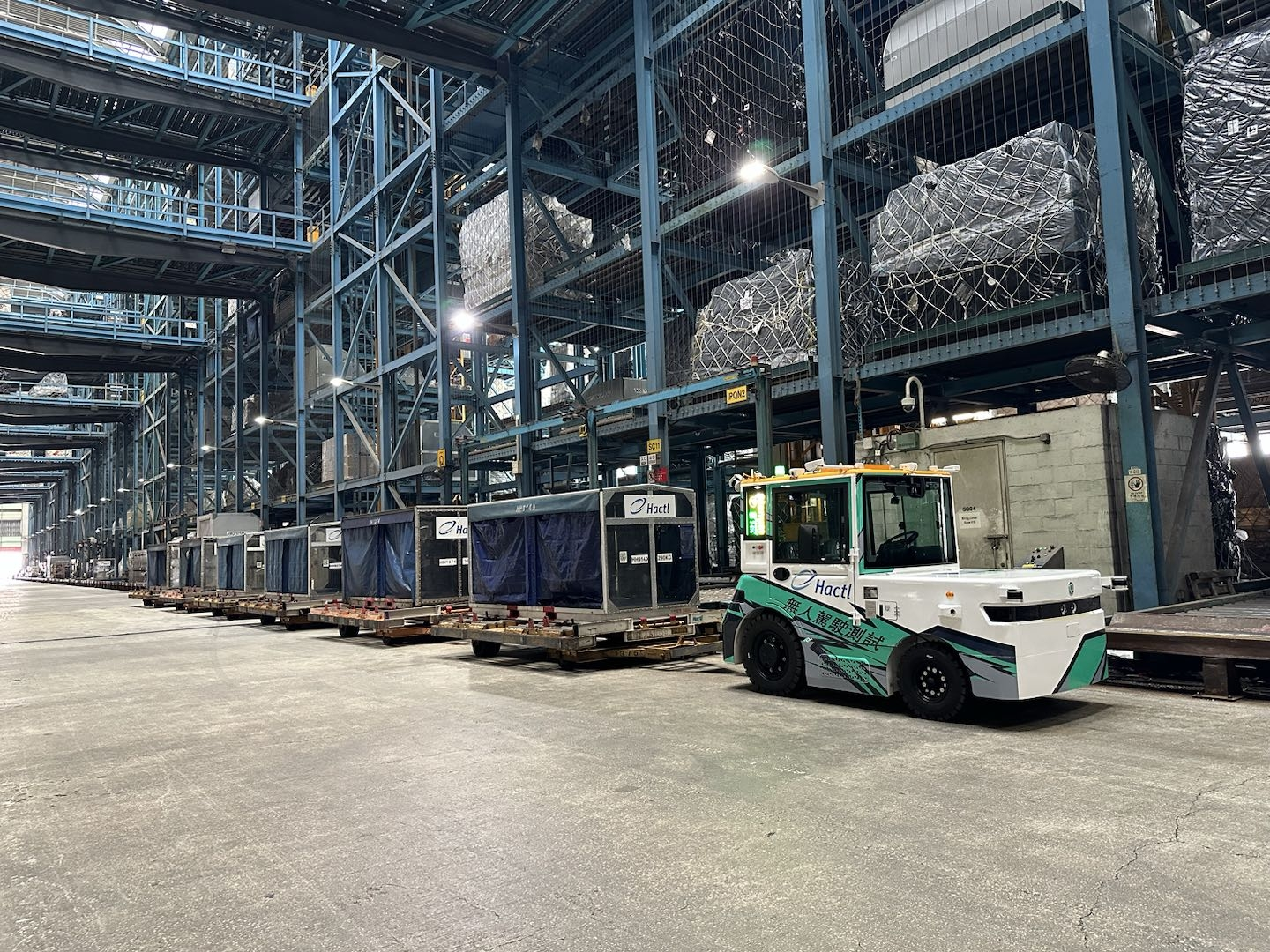 Autonomous electric vehicle transports cargo carts inside a modern industrial facility with automated warehouse systems and storage racks.