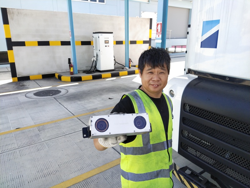 Technician showcases a vehicle-mounted sensor component beside an autonomous electric truck during equipment testing and inspection.